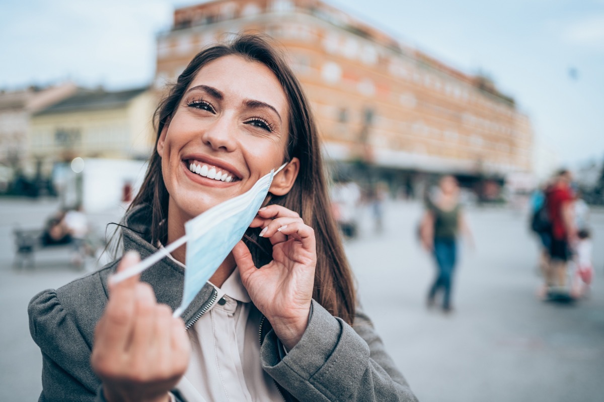 Beautiful happy young woman wearing protective medical facial mask for virus protection during Coronavirus/COVID-19 pandemic. Young woman putting/removing her mask outdoors in the city.