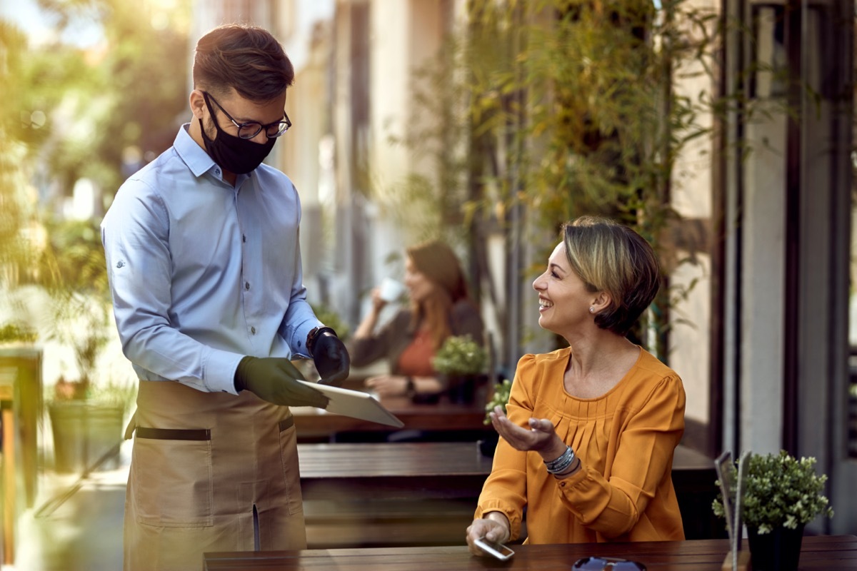 Happy waiter wearing protective face mask while showing menu on digital tablet to female guest in a cafe.