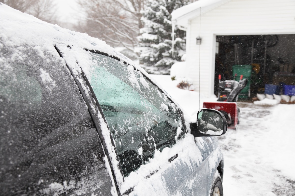 The snowblower in the background has just finished clearing the driveway while the brushed off car in the foreground warms up during a winter storm as heavy snow continues to fall. Selective focus on the car window.