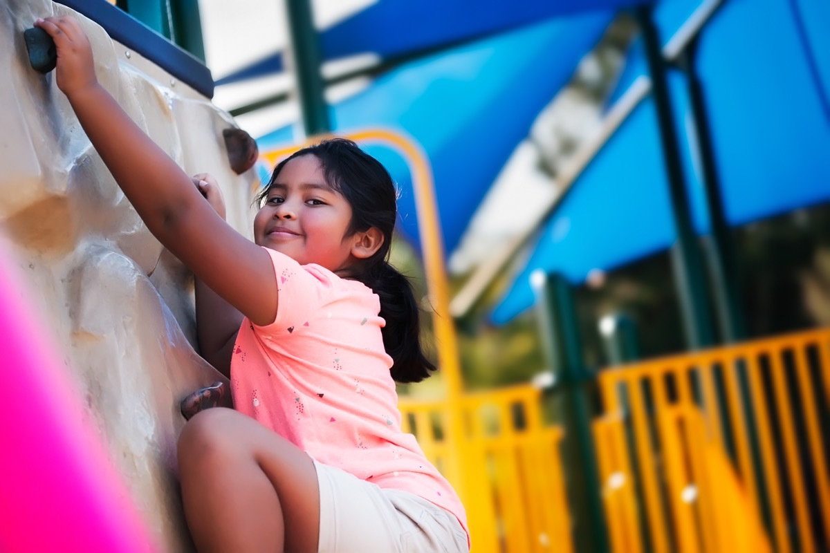 A smiling pre teen girl looking down while she reaches the top of a rock climbing wall gym.