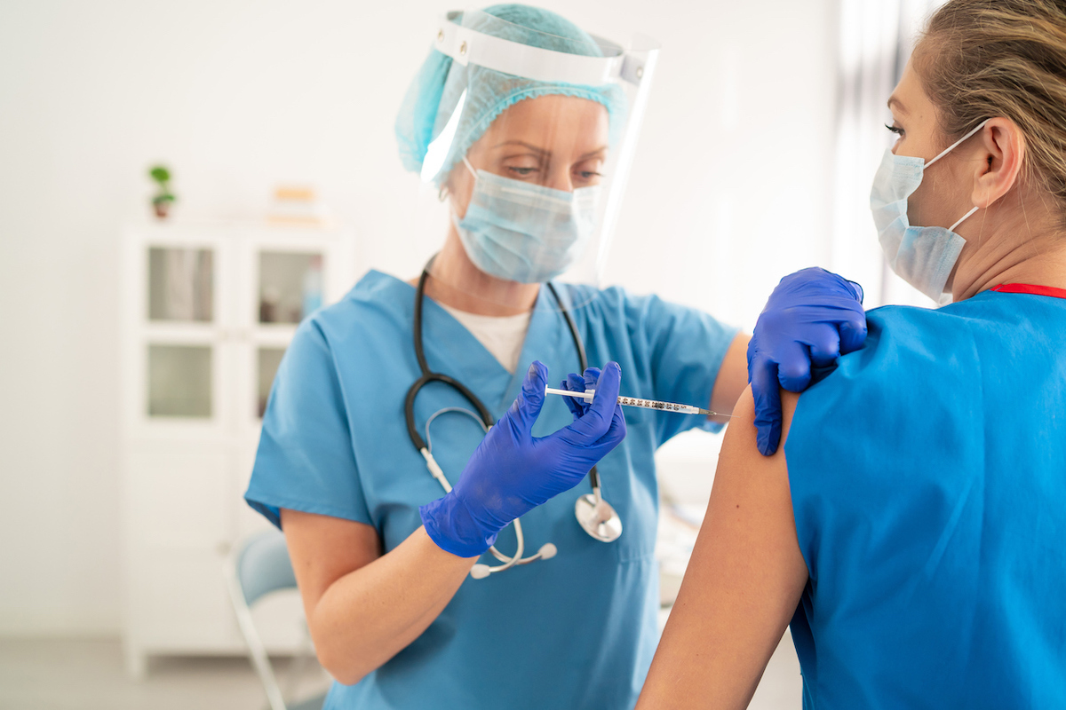 Doctor wearing protective face shield injecting COVID-19 vaccine to colleague