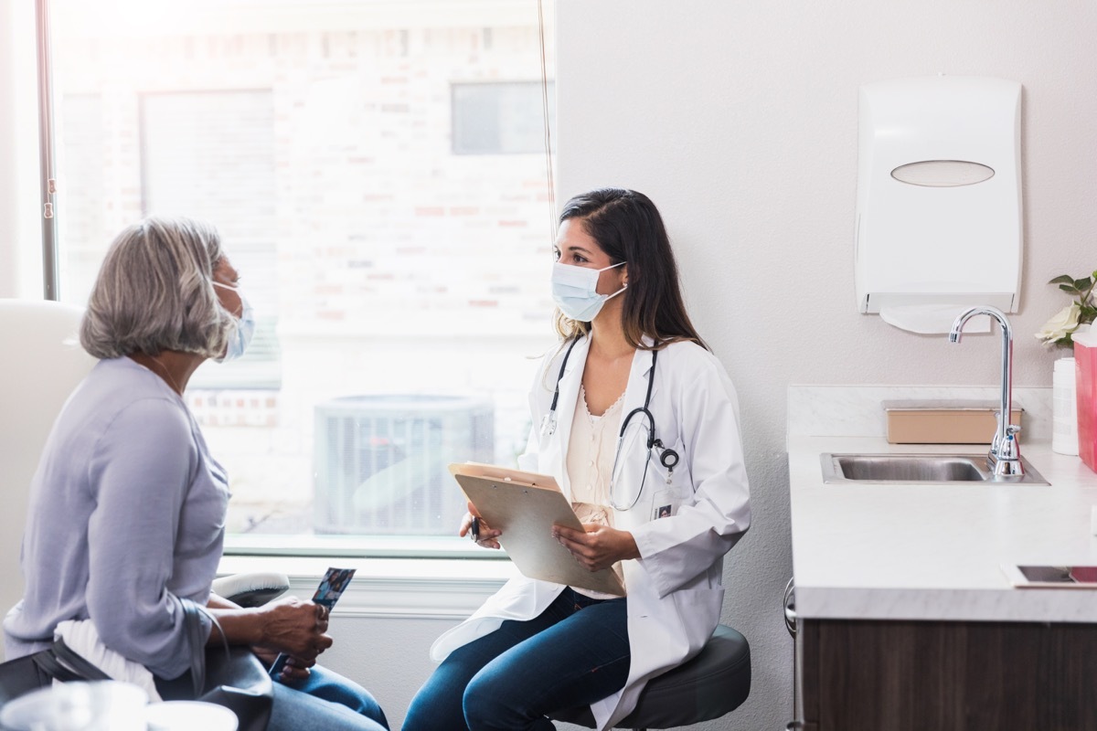 A senior woman listens as a female doctor gives a diagnosis. The doctor and patient are both wearing protective face masks as the patient is visiting the doctor's office during the COVID-19 crisis.